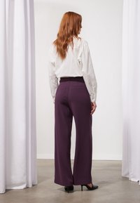 Paarse wijde broek met een gladde textuur, gecombineerd met een wit shirt met lange mouwen. De broek heeft achterzakken en een getailleerde pasvorm.