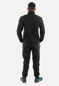 Zwart sportief trainingspak met een zipjack en broek met elastische tailleband. Bevat witte zijstrepen en contrasterende blauwe sneakers.