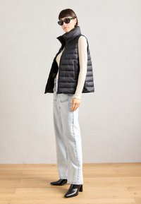 Gilet matelassé noir avec col montant, porté sur un haut à manches longues crème, associé à un jean en denim clair et des bottes noires à talons.