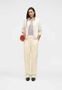 Γυναίκα που φοράει μπεζ πουλόβερ με σχέδια, γκρι μπλούζα, φαρδιά μπεζ παντελόνια, μαύρα παπούτσια και κρατάει μικρή κόκκινη τσάντα στον ώμο της.