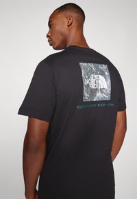 Camiseta de algodón negra con un gráfico cuadrado en la parte posterior que presenta el logo de "The North Face" y el texto "NEVER STOP EXPLORING" en color teal.