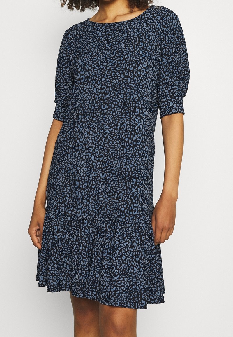 Femme portant une robe sombre longueur genoux avec un motif léopard bleu et des manches bouffantes aux coudes.