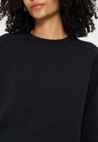 Personne aux cheveux bouclés portant un t-shirt noir uni à col rond, montrant le cou, les lèvres et une partie du visage sur fond blanc.
