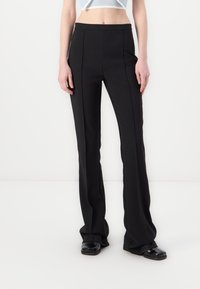 Pantalons noirs évasés avec une texture lisse, présentant des coutures avant et un design taille haute, associés à des chaussures noires plates.