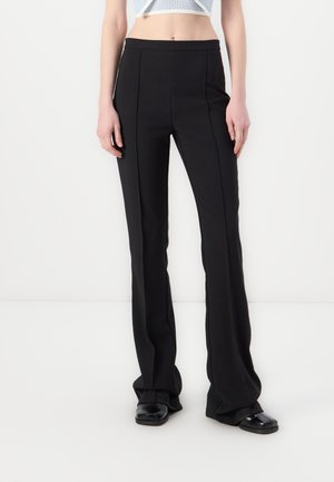 Pantaloni neri a palazzo con una texture liscia, caratterizzati da cuciture frontali e un design a vita alta, abbinati a scarpe nere flat.