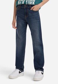 Άτομο που φοράει σκούρους μπλε ίσιους παντελόνι, τι-σάρτ «Νέα Υόρκη» σε τιρκουάζ και άσπρα αθλητικά παπούτσια Nike, στέκεται με το ένα χέρι στην τσέπη.