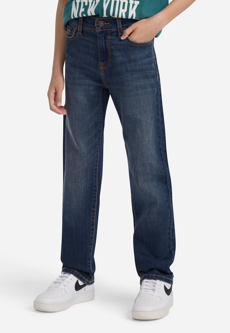 Άτομο που φοράει σκούρους μπλε ίσιους παντελόνι, τι-σάρτ «Νέα Υόρκη» σε τιρκουάζ και άσπρα αθλητικά παπούτσια Nike, στέκεται με το ένα χέρι στην τσέπη.