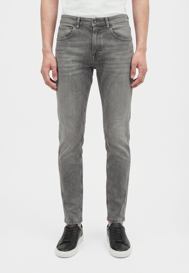 Jean gris slim porté par une personne avec les mains détendues sur les côtés, associé à des baskets noires à semelles blanches et une chemise blanche.