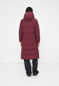 Geacă puffer maron, lungă, cu glugă, având cusături orizontale. Pură asupra unei perechi de blugi negri cu picior drept și adidași albi.
