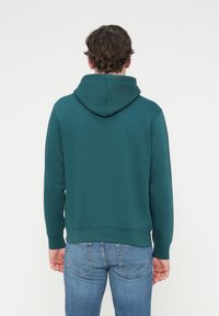 Φούτερ σε τυρκουάζ με κορδόνι στη κουκούλα, μακριά μανίκια και ριμπ μανσέτες. Λείο υλικό, φορεμένο με μπλε τζιν.