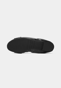 Semelle de chaussure noire avec une surface texturée antidérapante et un petit détail de boucle sur un fond clair uni.