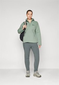 Žena u zelenoj jakni s patentnim zatvaračem, sivi hlačama i planinarskim cipelama, drži crnu trakicu od ruksaka, stoji pred jednostavnom bijelom pozadinom.