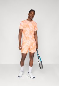 Herresports-outfit i lys orange med abstrakte mønstre, kortærmet skjorte og shorts. Inkluderer en tennisketsjer og hvide sneakers med blå accenter.