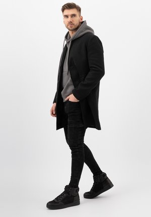 Cappotto nero sopra una felpa grigia, jeans neri attillati e sneakers alte nere. Il cappotto ha un design elegante e chiusure a bottoni.