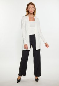 Witte lichte cardigan over een aangesloten wit topje, gecombineerd met zwarte wijde broek. Gestructureerde stof, open voorkant en lange mouwen.