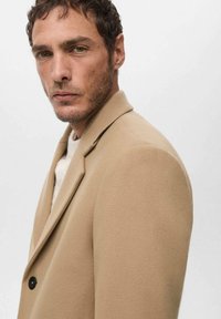 Blazer beige de mezcla de lana con solapa de muesca, acabado texturizado y detalle de botones negros. Corte slim, usado sobre un suéter ligero.