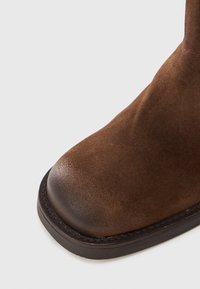 Bota de tobillo de ante marrón con punta redondeada, textura suave y suela oscura contrastante con costuras visibles alrededor del borde.