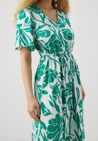 Robe fleurie verte et blanche en tissu léger, avec des manches courtes, un col en V, des détails de boutons et une taille élastique.