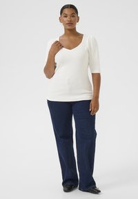 Kvinna med mörkt hår i en vit V-ringad topp, mörkblå jeans och svarta skor som står framför en ljus, enfärgad bakgrund.