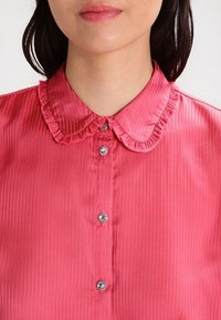 Rosa Satinbluse mit Rüschensaum, vertikal gestreiftem Muster und silbernen Knopfdetails an der Vorderseite. Weicher Stoff und taillierter Schnitt.