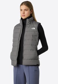 Harmaa puffaliivi, jossa on korkea kaulus ja etuvetoketju, nahanpintainen ompelutyö ja The North Face -logo vasemmassa rinnassa.