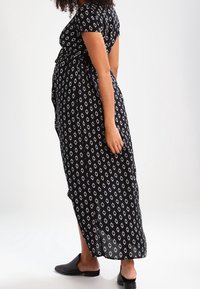 Robe maxi noire à manches courtes, motif en losanges blancs, taille cintrée et coupe ample. Associée à des chaussures noires à enfiler.