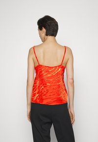 Camisola de seda vermelha com um padrão texturizado ondulado, alças finas ajustáveis e um design de costas em forma de U, combinada com calças pretas.
