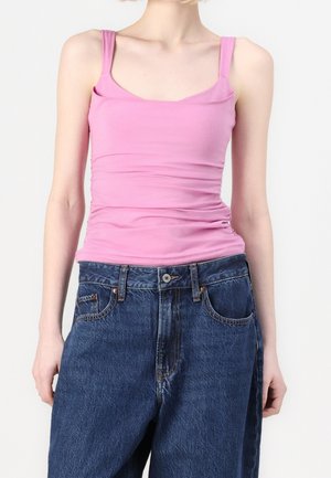 Osoba ubrana w dopasowany różowy top na ramiączkach w połączeniu z luźnymi ciemnoniebieskimi dżinsami na jednolitym tle.