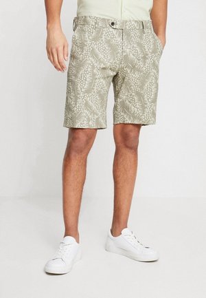Mand iført beige shorts med bladmotiv og hvide sneakers, står med hænderne delvist i lommerne på en ensfarvet lys baggrund.