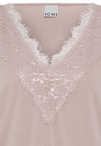 Blouse rose clair avec un col en V présentant une délicate bordure en dentelle florale et une étiquette de marque blanche "ICHI" visible au niveau du col.
