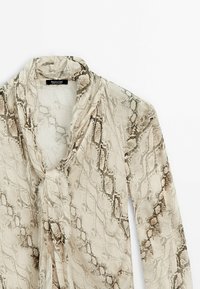 Blouse à manches longues beige clair avec un motif imprimé serpent marron et un col drapé, suspendue sur un fond blanc.