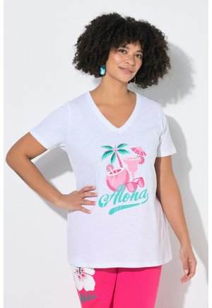 Frau mit lockigem Haar, die ein weißes V-Ausschnitt-T-Shirt mit Grafik von Palmen und Getränken trägt, rosa Hose mit weißen Hibiskusblumen und dem Schriftzug "Aloha".