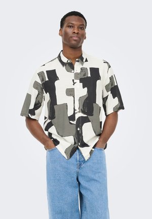 Hombre de pie con las manos en los bolsillos, vistiendo una camisa de manga corta abotonada con patrones abstractos en negro y gris, y jeans azul claro.