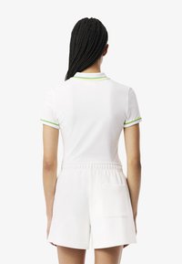 Polo bianco con accenti verdi e gialli sul colletto, abbinato a pantaloni corti bianchi con coulisse e una tasca sul retro. Texture liscia, design casual.