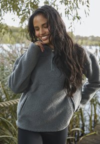 Pullover in pile grigio con mezza zip, maniche lunghe, due tasche laterali e finitura testurizzata, ambientato su uno sfondo naturale.