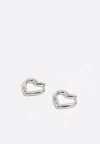 Pendientes en forma de corazón de plata con una superficie lisa y brillante; pequeña grabación de la marca visible. Cada pendiente presenta un diseño recortado distintivo.