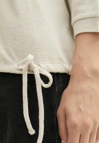 Világos bézs ing, puha tapintással, kötött zsinórral a hemnél. Kéz pihen egy fekete ruhán, denimszerű textúrával.