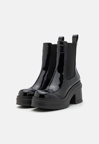 Botas Chelsea de charol negro con tacones gruesos, paneles laterales elásticos, punteras redondeadas y acentos de logo en la parte delantera y trasera.