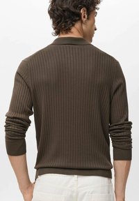 Man in een bruine geribbelde trui met lange mouwen en kraag, gezien vanaf de achterkant, gecombineerd met witte broek.