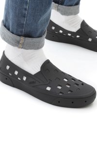 Osoba oblečená v bílých ponožkách a černých perforovaných slip-on teniskách Vans, s vyhrnutými modrými džíny na bílém pozadí.
