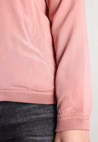 Jasnoróżowa, lekka kurtka z długimi rękawami, prążkowanymi mankietami i zaokrąglonym dołem. Posiada boczną kieszeń o gładkiej fakturze.