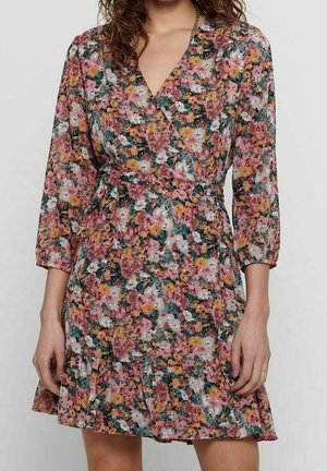 Femme portant une robe fleurie aux manches longues et longueur genou, avec des couleurs rose, orange, blanc et vert, et un ourlet à volants.