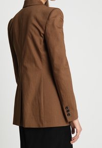 Nainen, jolla on päällään hyvin istuva ruskea bleiseri, jossa on kolme mustaa nappia hihassa, näkyy selkäpuolelta yksinkertaista taustaa vasten.