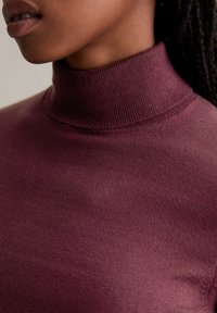 Burgundi turtleneck pulóver sima textúrával és bordás gallérral, testhezálló fazonnal, kiemelve a dekoltázst és a puha anyagot.