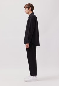 Schwarzes Langarmhemd mit spitzem Kragen, Brusttasche; kombiniert mit schwarzer Hose und weißen Sneakers; glatte Stoffstruktur.