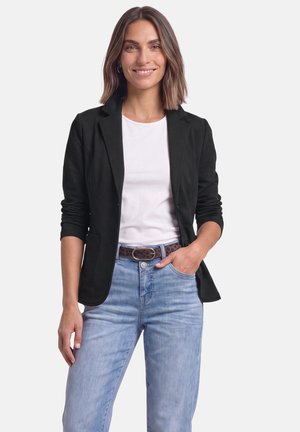 Donna con capelli castani fino alle spalle, che indossa un blazer nero sopra una camicia bianca e jeans azzurri chiari, sorridente con una mano in tasca.