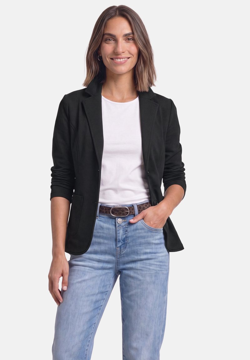 Femme aux cheveux bruns mi-longs, portant un blazer noir sur une chemise blanche et un jean bleu clair, souriant avec une main dans la poche.