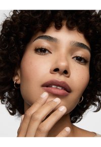 Cheveux noirs bouclés, peau brun clair, portant de petites créoles en or. Lèvres d'un mauve doux, vernis à ongles beige clair, main touchant doucement le visage.