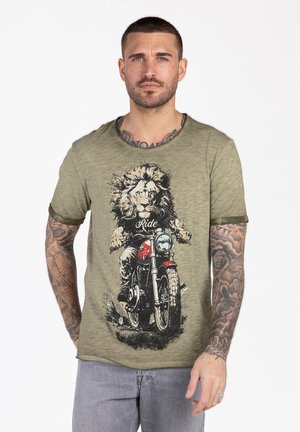 Uomo con tatuaggi che indossa una maglietta verde oliva con una grafica di un leone che guida una moto rossa e pantaloni grigi, in piedi su sfondo bianco.