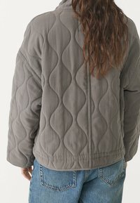 Een persoon in een grijze gewatteerde jas met golvende stikpatronen en losse blauwe spijkerbroek, gezien vanaf de achterkant met lang bruin haar zichtbaar.